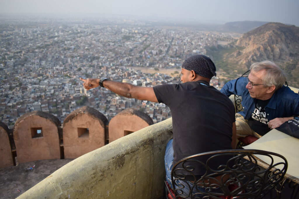 A local guide pointing out views of Jaipur city from a hilltop fort, while a foreign tourist listens attentively, with a panoramic cityscape in the background.