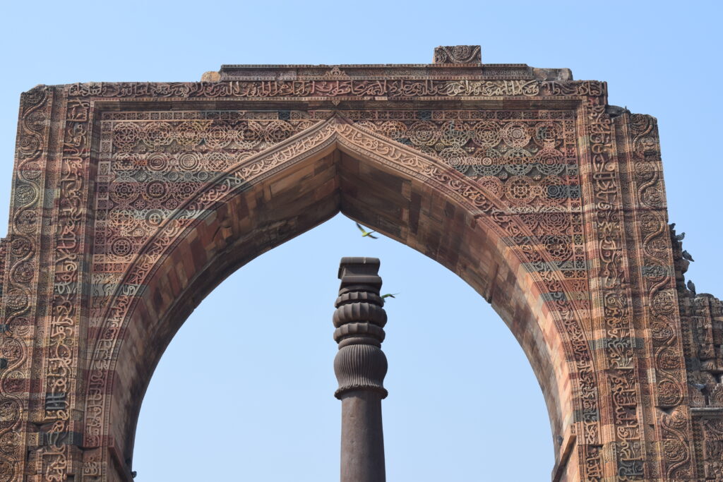 Intricately carved sandstone arch of the Qutub Minar complex framing the ancient Iron Pillar of Delhi against a clear blue sky.
