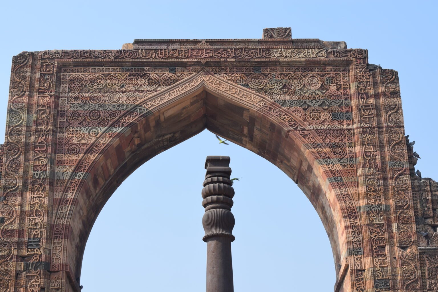 Intricately carved sandstone arch of the Qutub Minar complex framing the ancient Iron Pillar of Delhi against a clear blue sky.