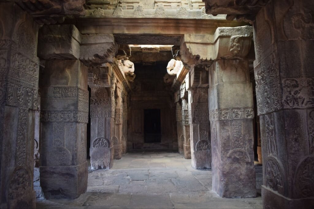 A view looking down a narrow, dimly lit passage within an ancient stone temple, featuring intricately carved stone pillars and ceiling beams.