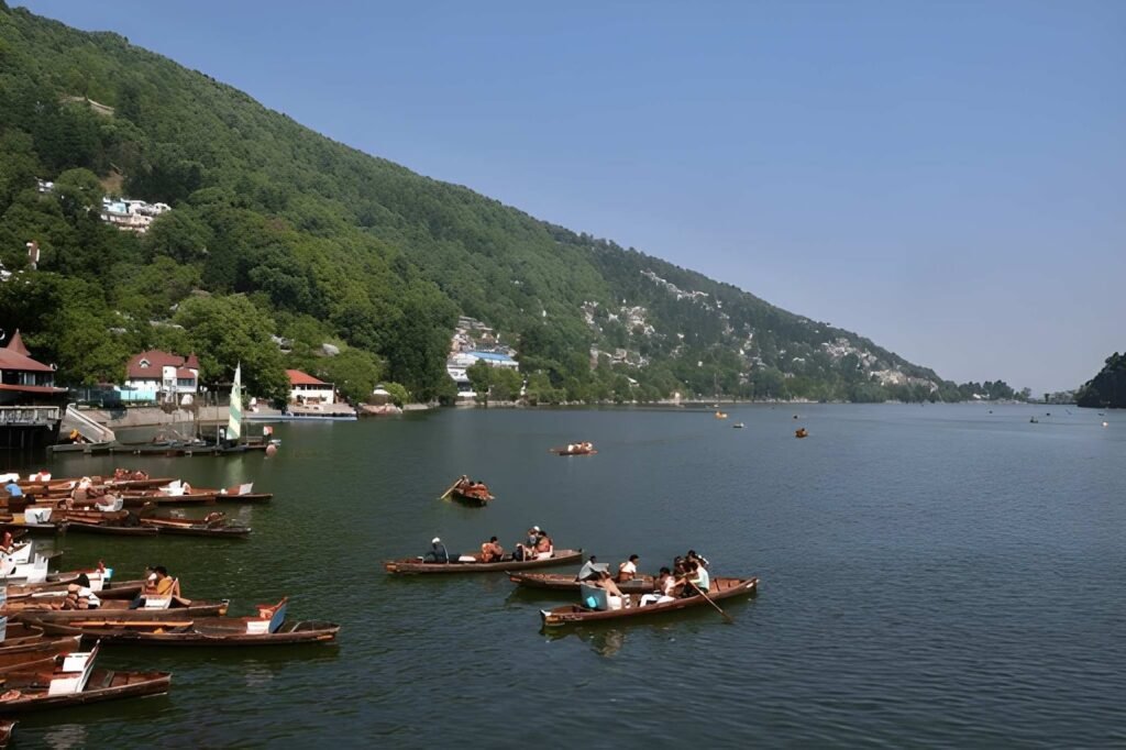 A scenic view of the crescent-shaped Naini Lake in Nainital, India, surrounded by lush green hills, with many wooden rowboats carrying tourists on the water and docked near the shore.