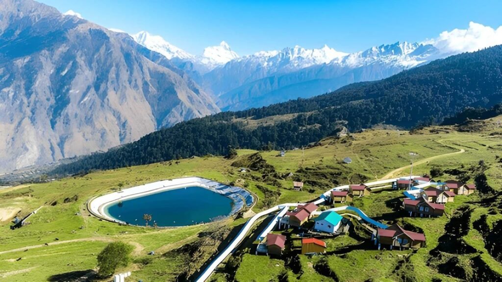 An aerial view of the Auli Artificial Lake, one of the highest man-made lakes in the world, surrounded by lush green meadows and small resort buildings.