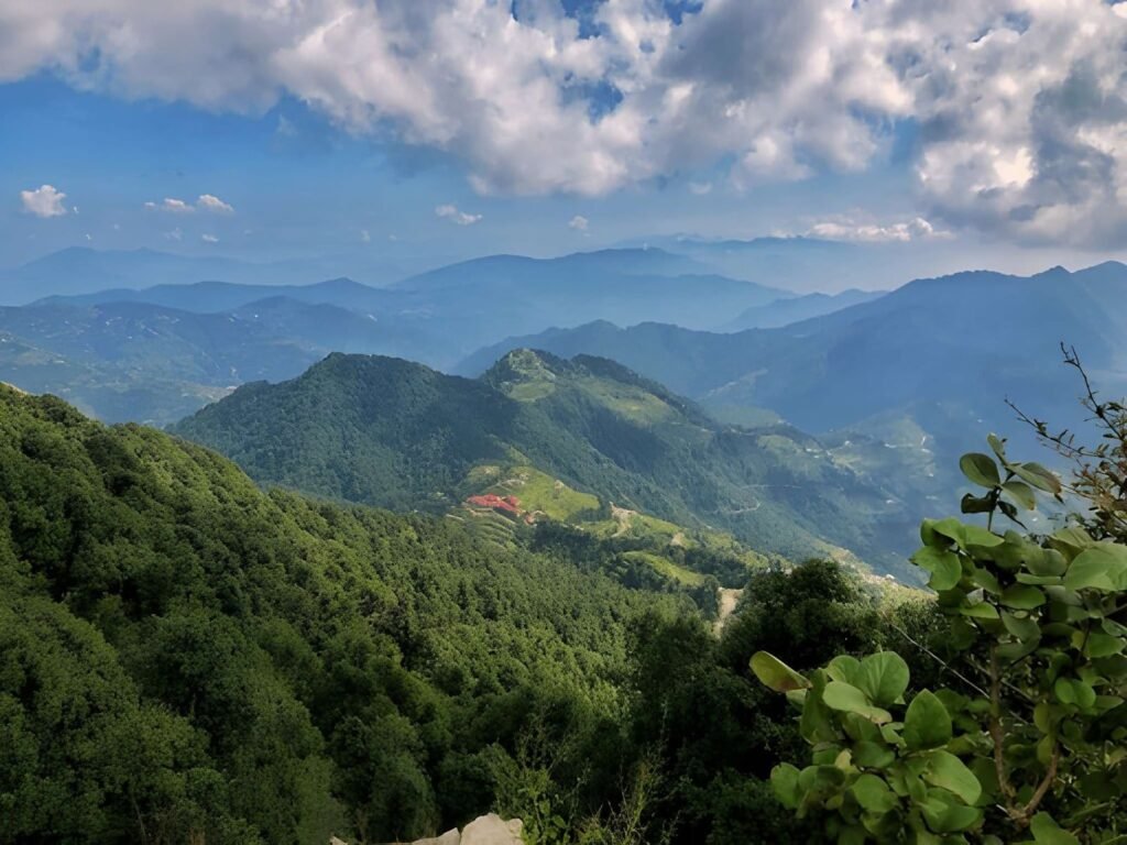 A view of lush green, forested mountains and valleys under a partly cloudy blue sky, likely in the Kanatal region of Uttarakhand, India.