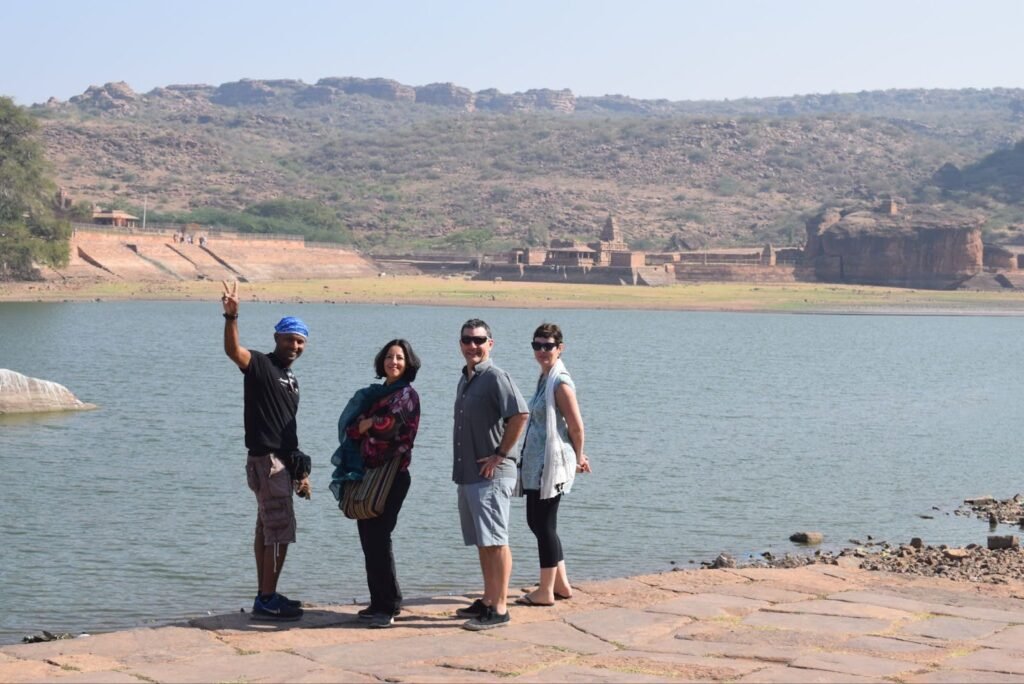 A group of four people standing beside a large body of water, with rocky hills and an ancient temple structure visible in the background.
