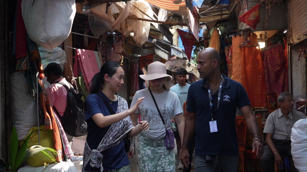 A bustling market scene in Mumbai, featuring Reality Tours & Travel guides wearing navy blue polo shirts as tehy lead a group of international tourists through a vibrant stall filled with colorful textilies.