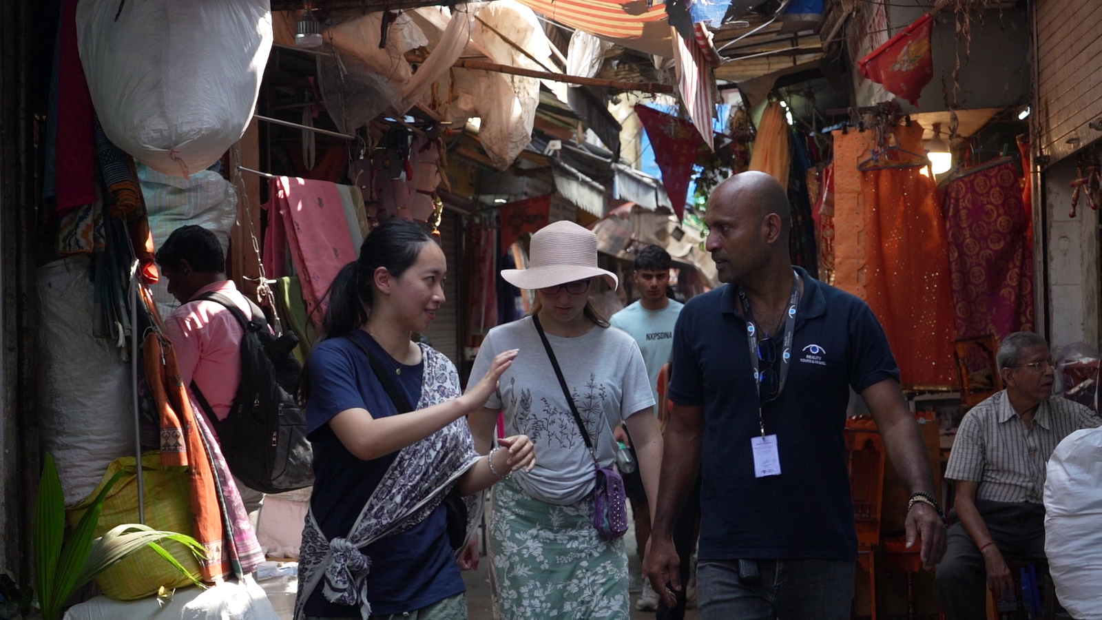 A bustling market scene in Mumbai, featuring Reality Tours & Travel guides wearing navy blue polo shirts as tehy lead a group of international tourists through a vibrant stall filled with colorful textilies.