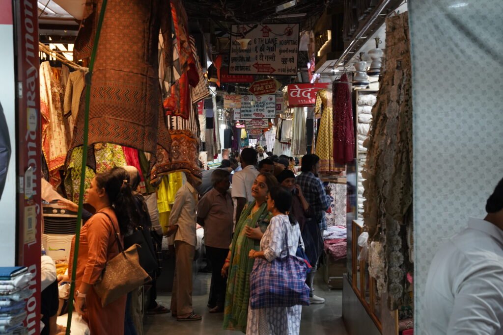A narrow but bustling lane inside Mumbai's Mangaldas Market, crowded with shops displaying stacks of colorful ethnic fabrics and hanging garments.