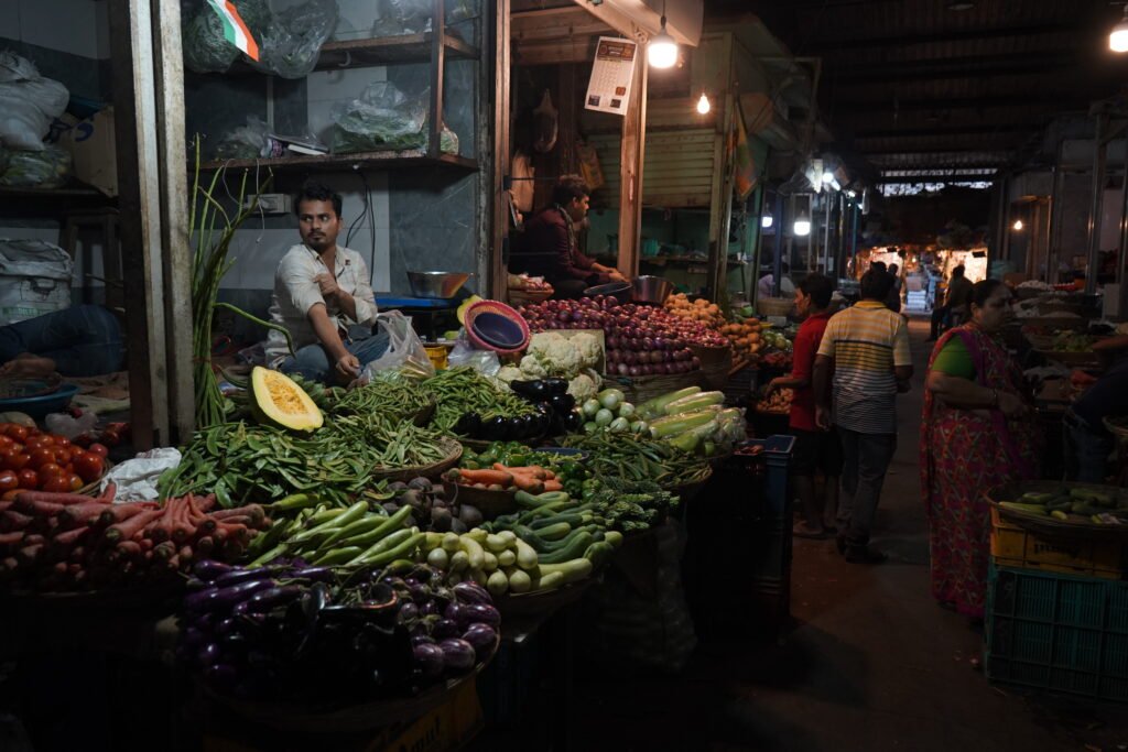 A vibrant night scene at Null Bazaar in Mumbai, showing a street vendor seated behind a large, colorful display of fresh vegetables like carrots, eggplants, and leafy greens under warm market lights.
