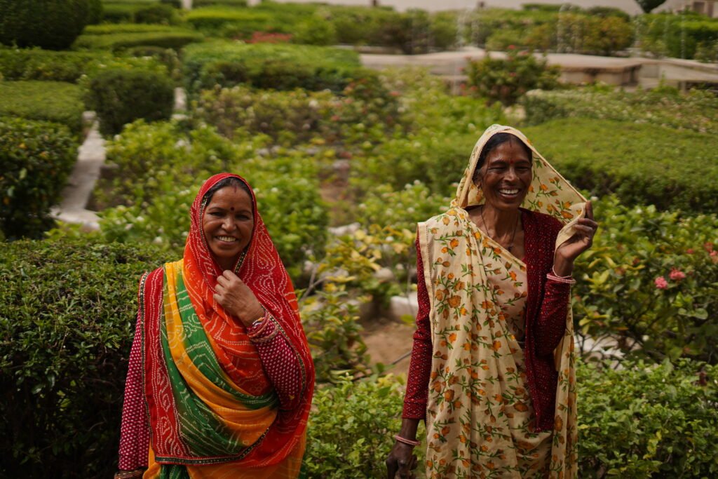 Two Indian women smiling in traditional attire, standing in a lush, landscaped garden setting.