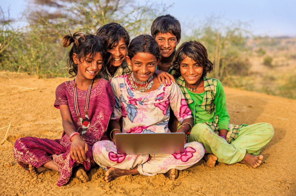 A group of happy Indian children sitting outdoors looking at a laptop, representing digital India.